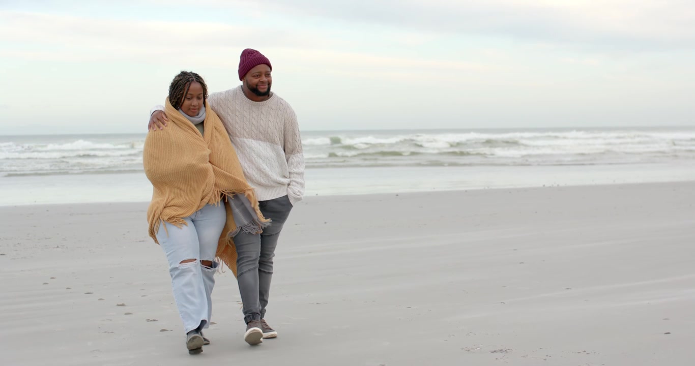 African American couple strolling slowly on beach, blanket fluttering in wind, man wearing beanie