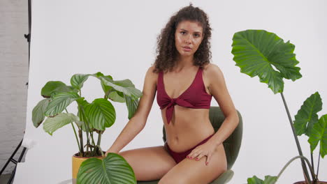 modelo bronceada y de cabello rizado oscuro posando en una silla con un traje de bikini rojo, rodeada de plantas, filmada en un estudio fotográfico