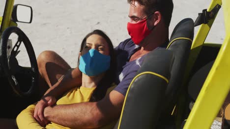 feliz pareja caucásica con máscaras faciales sentada en un buggy de playa