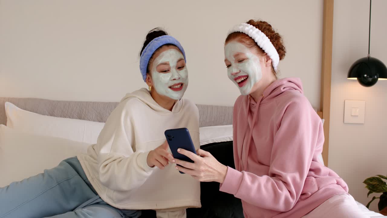 Diverse amiche sedute sul letto che indossano maschere e fasce per capelli, che tengono in mano un telefono per divertirsi, ridendo