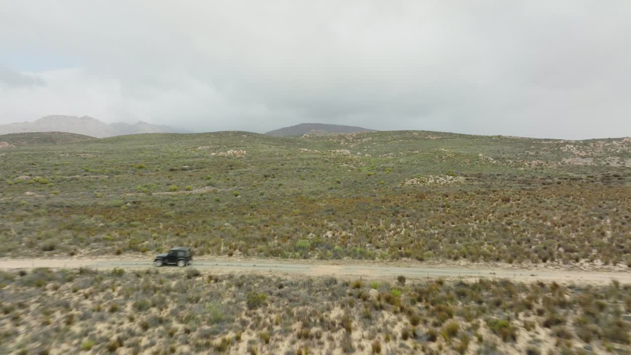 드론이 바위 지형 근처를 날아가서 검은색 오프로드 차량이 운전하는 도로를 가로질러 남아프리카 공화국의 세더버그 야생 지역 - 배경에는 바위 산을 볼 수 있습니다.