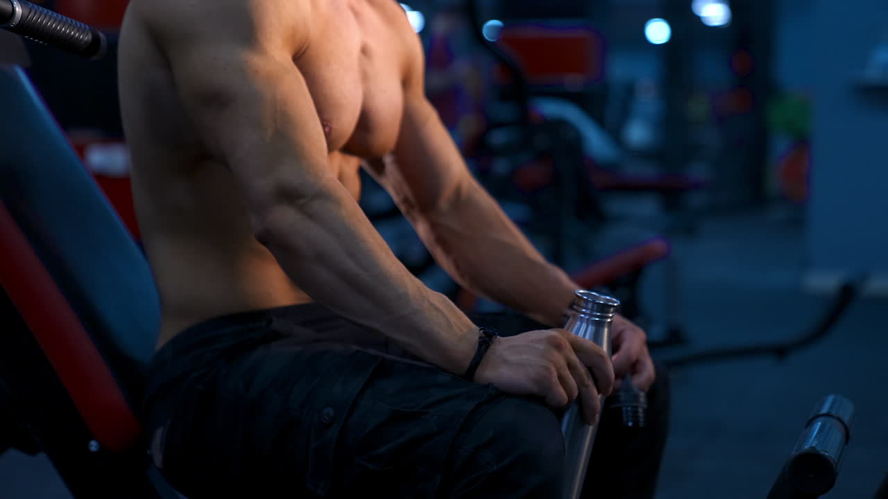 Rest in sports center. Shirtless man having break during workout. Athletic man sitting on a simulator with a flask of water. Close-up.