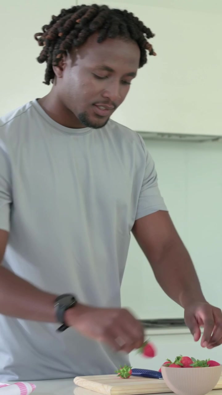 Vertical video: Adjusting towel, man in gray tee, watch trimming strawberry in kitchen for blender