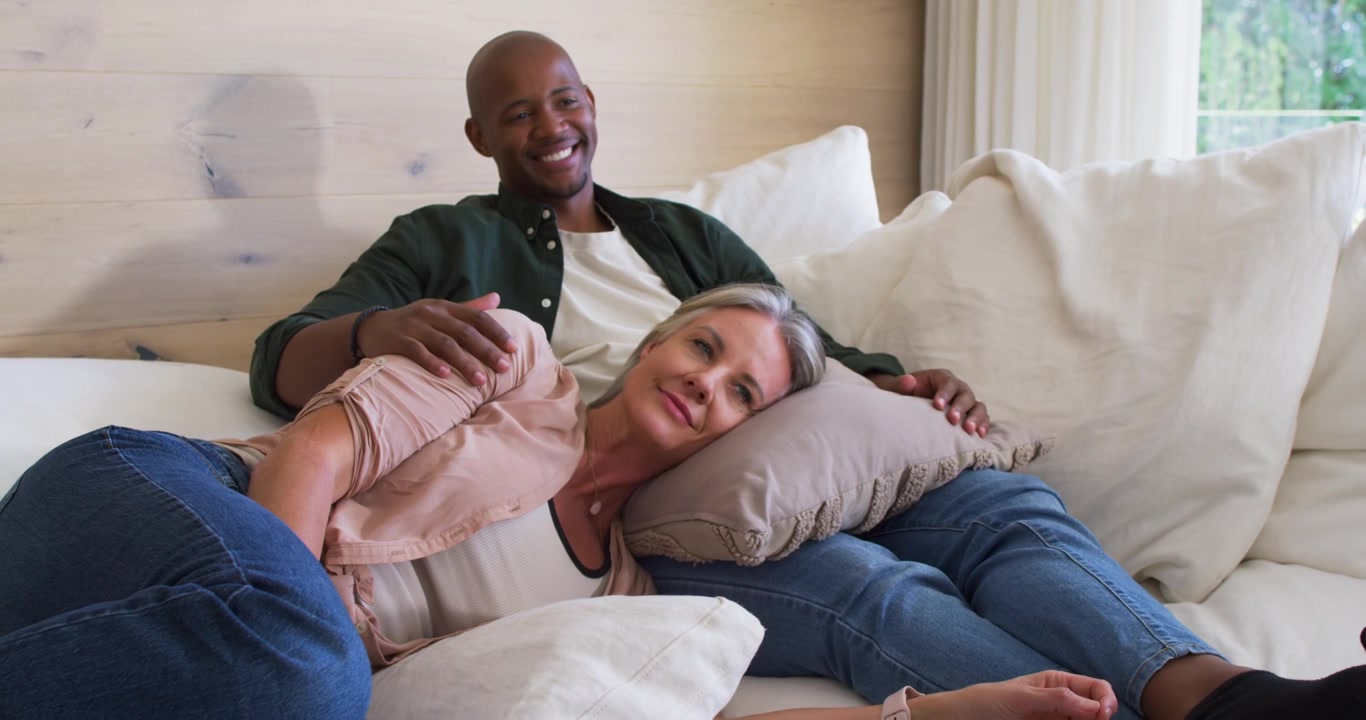 Mangfoldig par deler et rolig øyeblikk på sofaen hjemme, smiler, justerer pute og sjekker klokken.