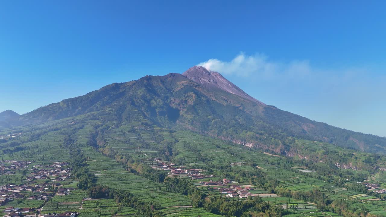 Aerial view of Mount Merapi volcano emitting smoke above lush green farmland under a clear blue sky
