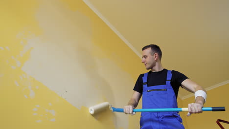 painter man at work painting a wall with a roller. worker in blue overalls doing renovation of a room. offering professional painting services. view from below.