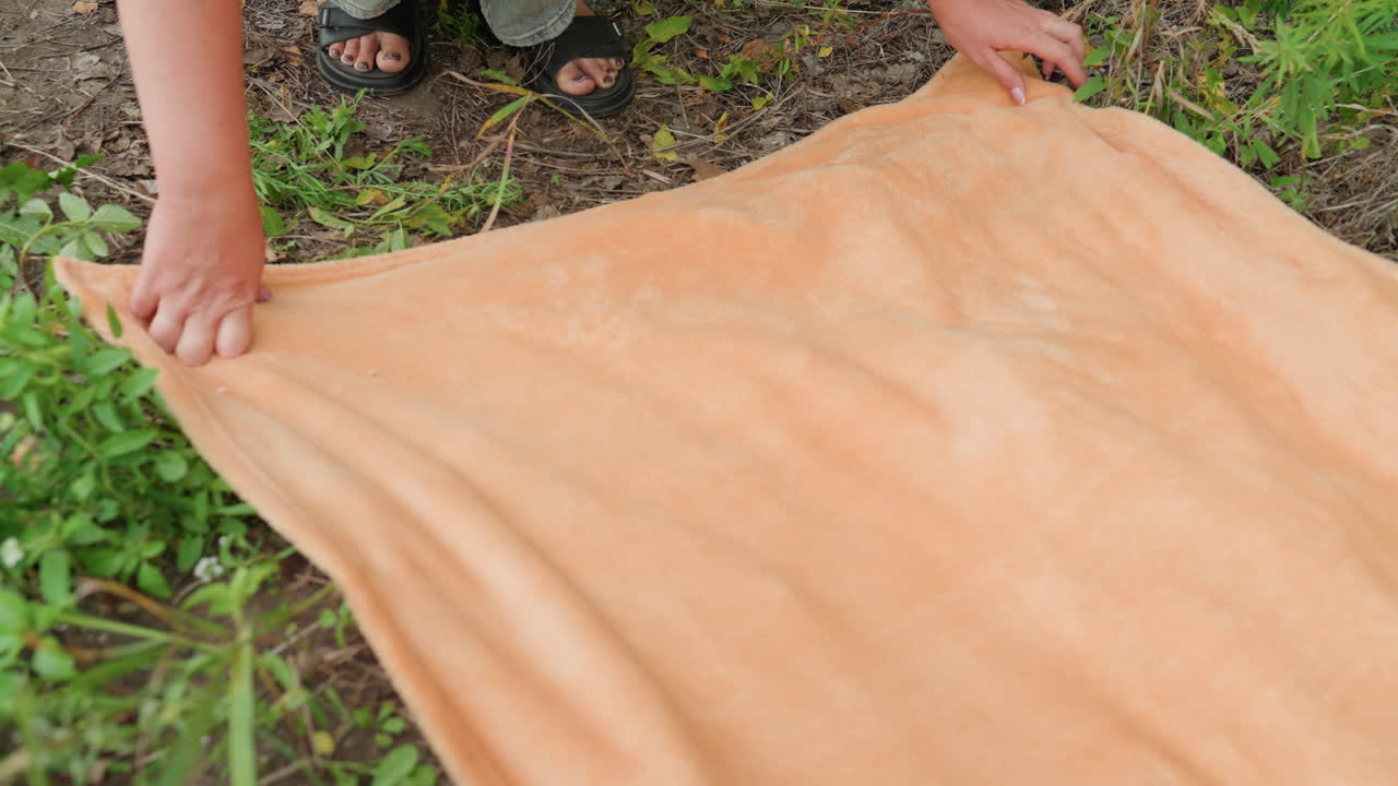 Child and parent organize blanket on grass outdoors, Family arranging picnic blanket together outside in sunlight, Parents and child carefully spread out blanket for picnicking outdoors together