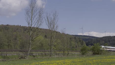 Tren-De-Alta-Velocidad-Acelerando-A-Través-De-Un-Vibrante-Paisaje-Rural-Con-Turbinas-Eólicas-En-El-Fondo,-Día-Soleado