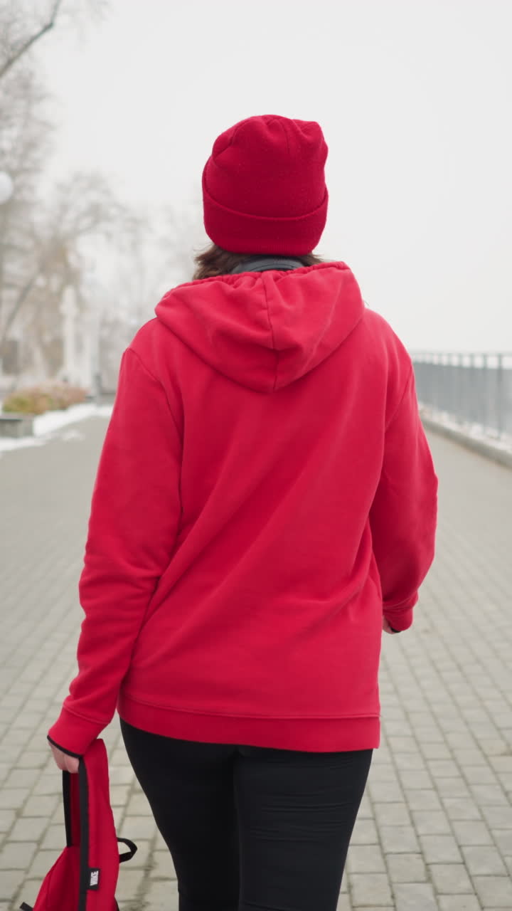 Back view of woman wearing red jacket walking along interlocked pathway holding bag in left hand near iron railing in serene park with snowy ground benches and trees in foggy winter atmosphere