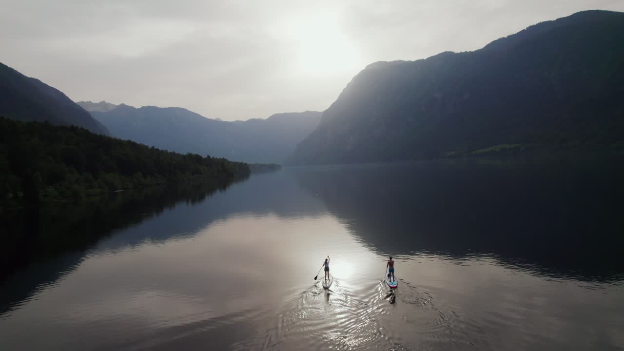 Drone shot over Lake Bohinj in Slovenia as a couple does stand up paddling with mountains in the backdrop during the evening