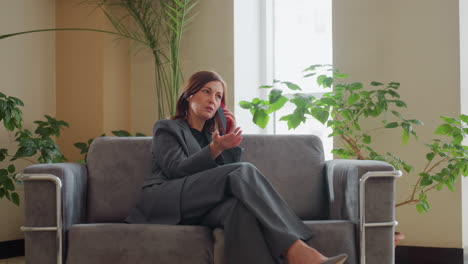 serious businesswoman in formal suit sitting on sofa holding smartphone to ear, engaged in intense phone conversation in bright modern office lobby with potted plants and large windows around her
