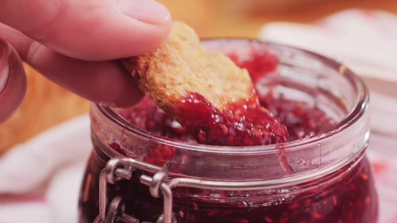 Hand dipping a biscuit into a jar of raspberry jam at breakfast
