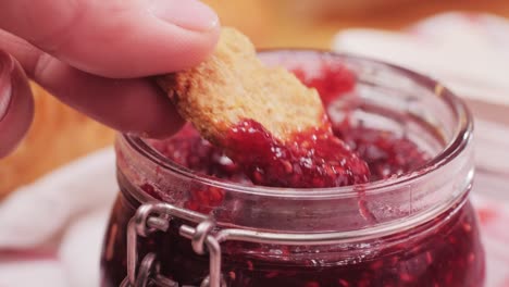 hand dipping a biscuit into a jar of raspberry jam at breakfast