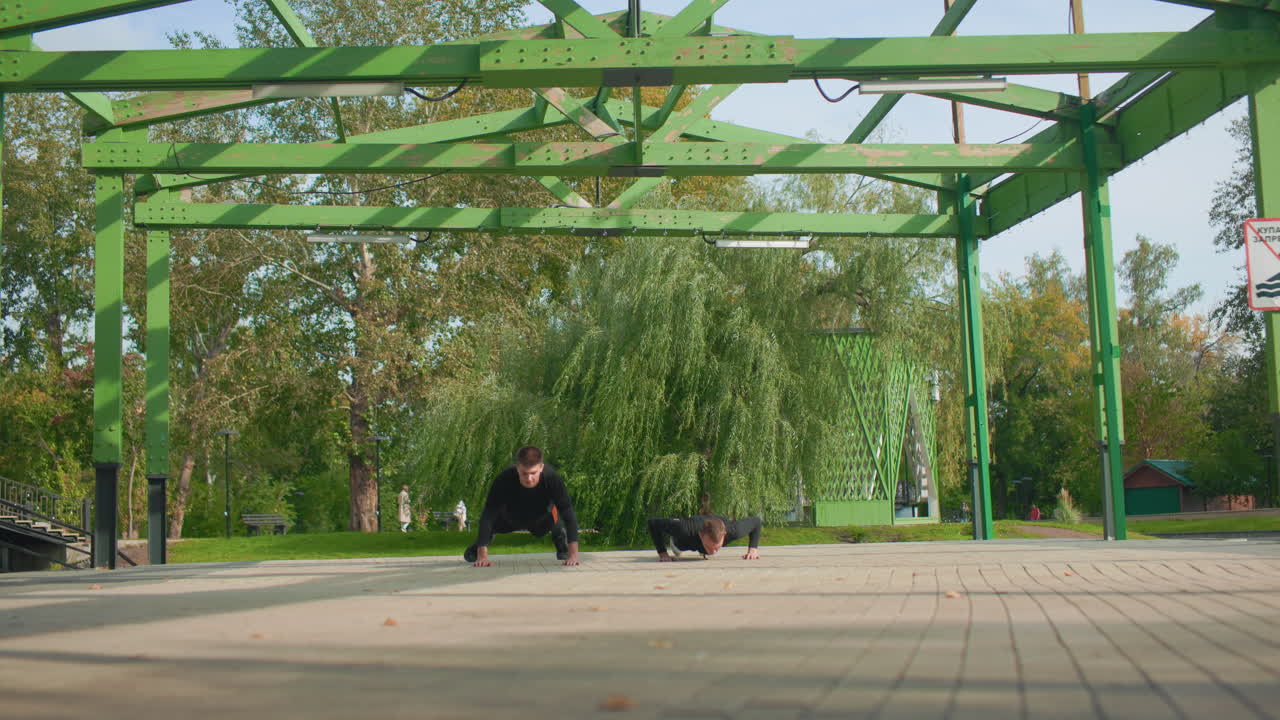 Deux athlètes faisant des pompes sous une pergola, plan large en contre-plongée d’un entraînement synchronisé au poids du corps sur une place pavée sous une pergola métallique verte dans un parc urbain, la lumière du soleil matinale filtrant à travers les feuilles