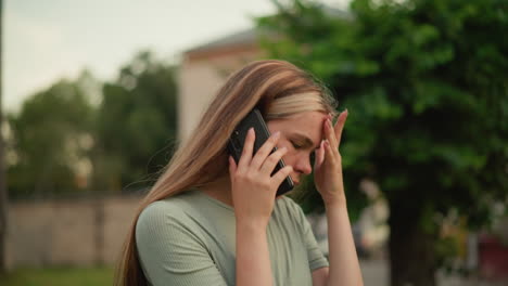 primer plano de una mujer joven que parece agotada durante una llamada telefónica, sosteniendo su mano izquierda en su frente en frustración, el fondo muestra vegetación borrosa y un edificio