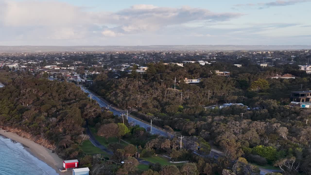 Drone glides over Rye beach at dusk, showcasing bathing boxes, coastal road, and lush greenery
