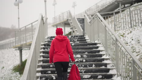 back view of woman in red hoodie walking up snowy staircase while carrying red backpack. iron railings line the stairs, surrounded by snowy landscape, with light poles adding to the wintry atmosphere