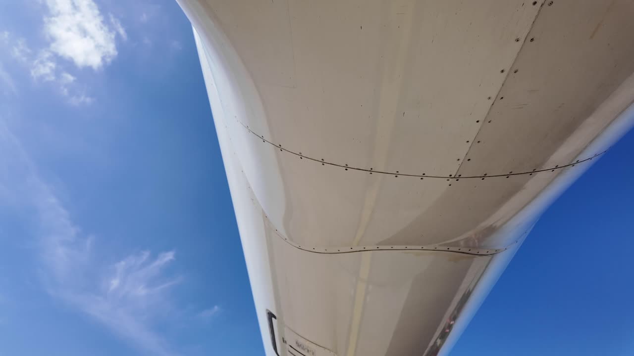 External view from the rear part of the lower part of a white color fuselage medium size jet, tilting down. Sunny day with a blue sky. 4K
