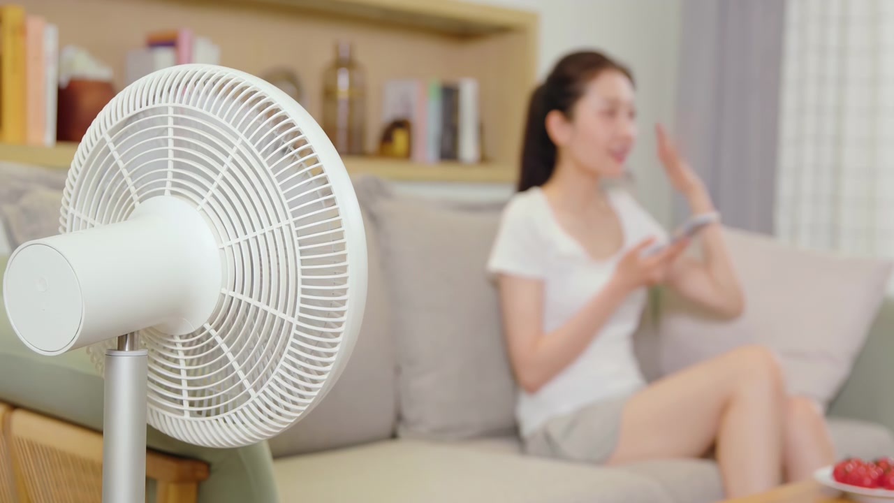 Woman relaxing sitting on cozy couch, using a smartphone while cooling down in front of electric fan at home during summer hot weather apartment. Female trying to fight the heat staying hydrated