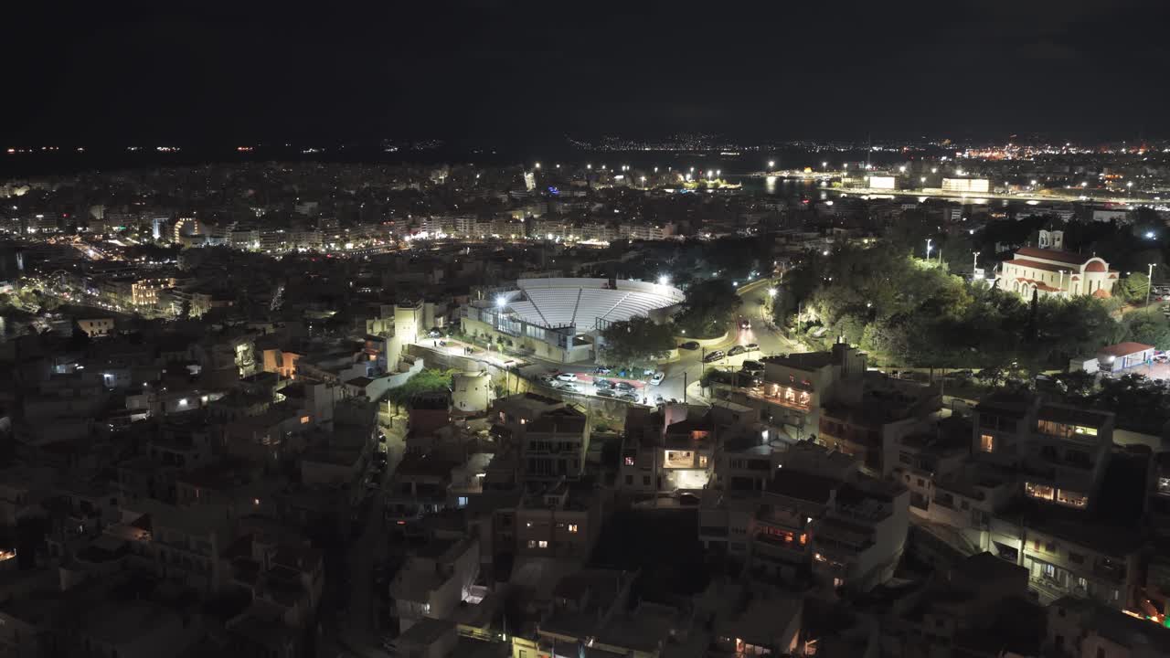 Splendide riprese aeree che catturano il paesaggio urbano illuminato del Pireo di notte, mostrando l'iconico teatro comunale Veakio, le strade animate e il porto illuminato in lontananza.