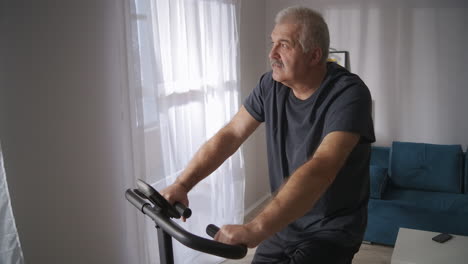 Un-Hombre-De-Mediana-Edad-Está-Entrenando-En-Casa-Girando-Pedales-De-Bicicleta-Estática-En-La-Sala-De-Estar-Con-Un-Estilo-De-Vida-Saludable-Y-Manteniendo-Una-Buena-Condición-Física-A-Mediana-Edad