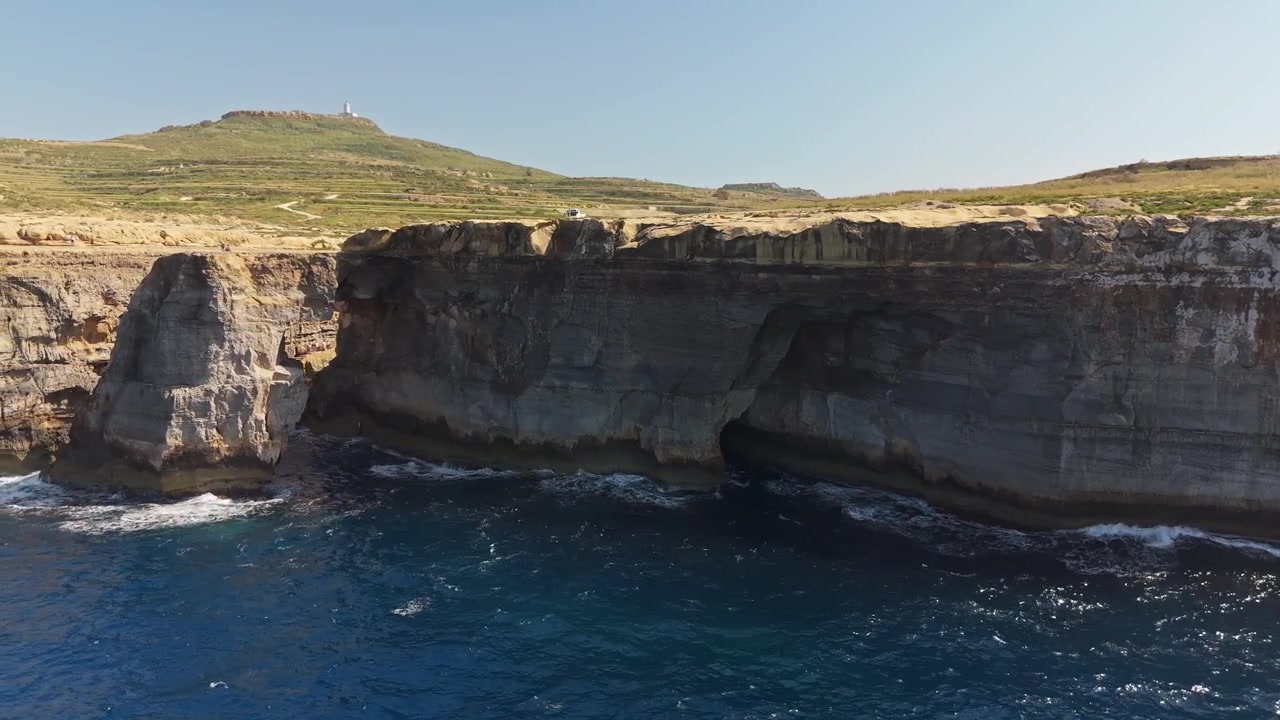 Wied il-Mielaħ in Gozo, Malta, natural rock arch and coastal cliffs above blue Mediterranean water, with visible stratified layers and waves along the rugged shoreline in aerial view.
