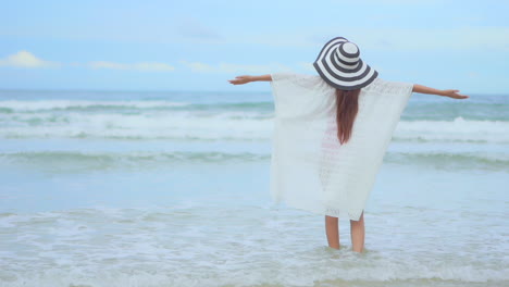 여름에 sundress와 줄무늬 모자를 쓰고 물가 해변에 서있는 아시아 여자