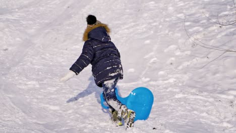 children on snow in winter. little boy rising up the hill with his plastic plate outdoors. joyful childhood during winter vacation. slow motion.