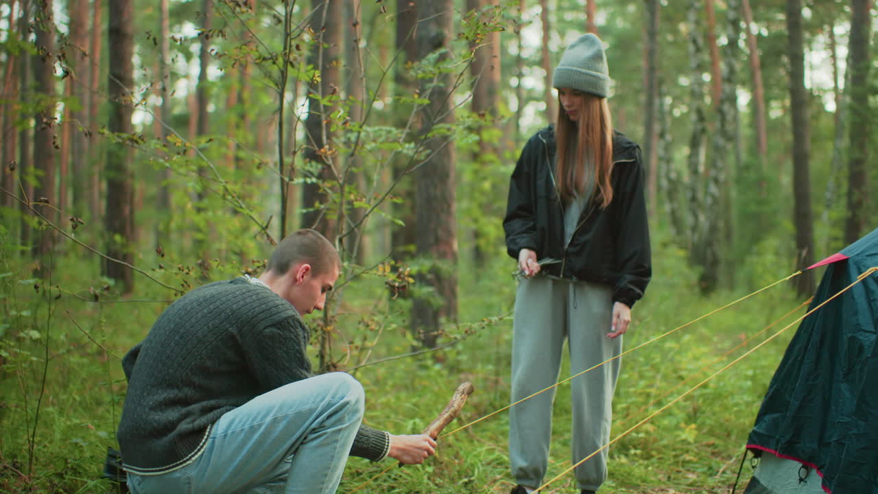 Young woman in casual outfit watches attentively as man crouches to secure yellow tent rope into forest ground, surrounded by trees and greenery during outdoor camping activity