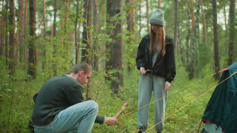 young woman in casual outfit watches attentively as man crouches to secure yellow tent rope into forest ground, surrounded by trees and greenery during outdoor camping activity