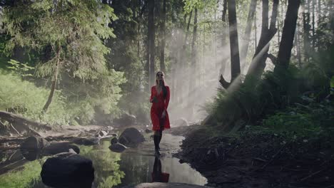 enchanted forest stroll: a woman in a vibrant red dress walking along a riverbed through a lush, fairy-tale like forest amid the mystical morning mist and fog