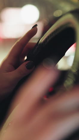 vista en primera persona de la mano de una mujer con uñas pintadas de negro tocando un volante. la escena es capturada por la noche con luces de la ciudad coloridas y borrosas en el fondo
