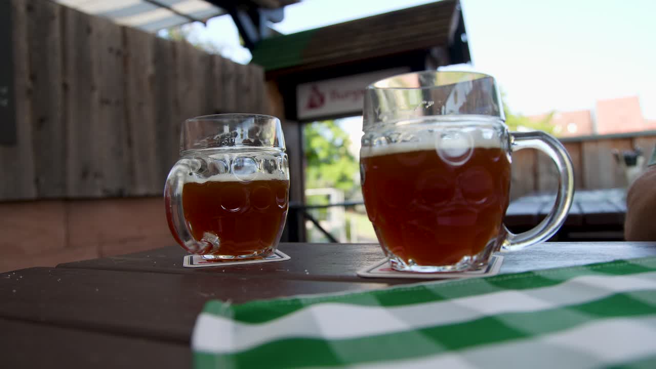 Person grabs glass beer mug on wooden table in sunny outdoor beer garden, natural daylight