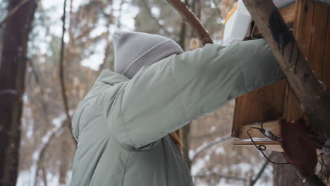 woman in gray winter jacket and knit beanie observing wooden bird feeder in snowy forest, focused expression, close-up side view, quiet moment in nature, serene winter woodland atmosphere