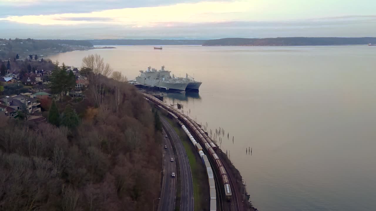 Long Train On The Railroad By The Bay In Washington, USA With Ships Docked Over The Calm Ocean Near The Shore On A Sunset - Aerial Shot