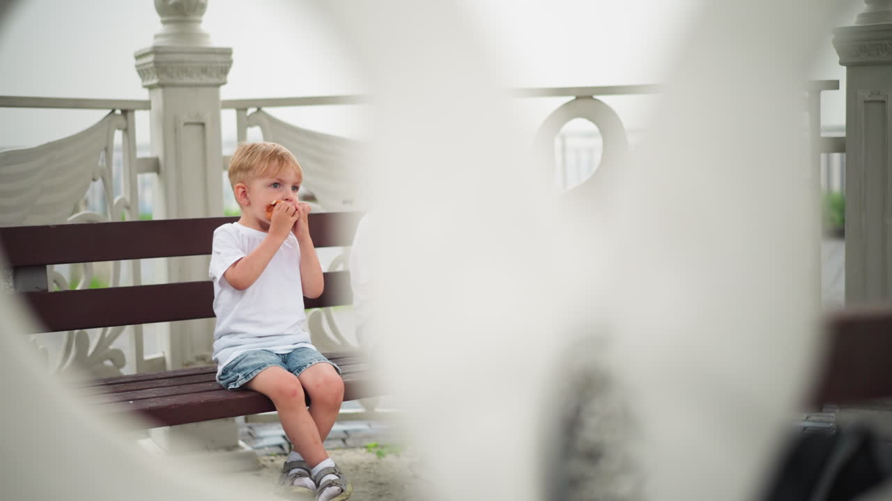 маленький мальчик наслаждается колбасами, сидя на скамейке, а рядом с ним сидит старший мальчик, который, по-видимому, сосредоточен на чем-то другом.