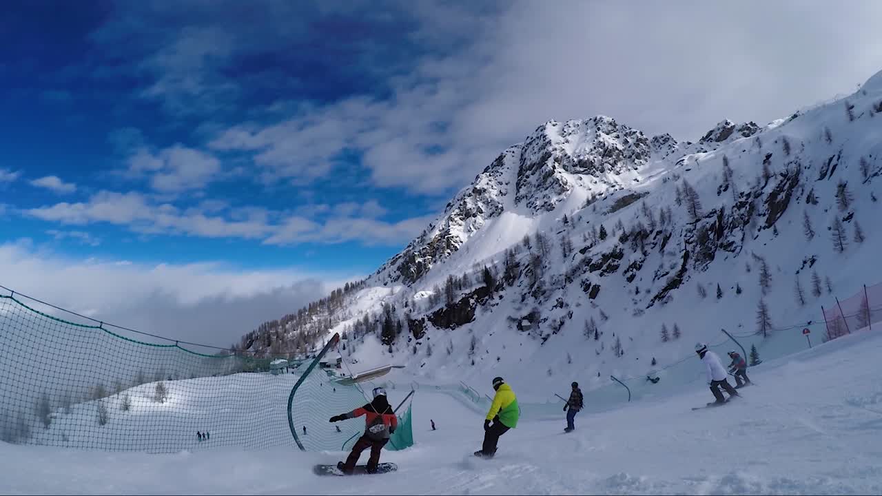 Following snowboarders going down a ski slope with nets on sides and a snowy mountain in the background with partly cloudy blue sky
