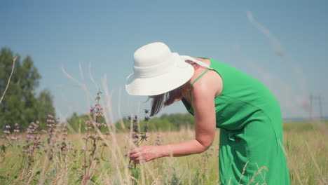 side view of woman bending forward in grassy meadow wearing green dress and white hat to smell lavender blooms near treeline under clear sky with distant power lines visible soft breeze stirring
