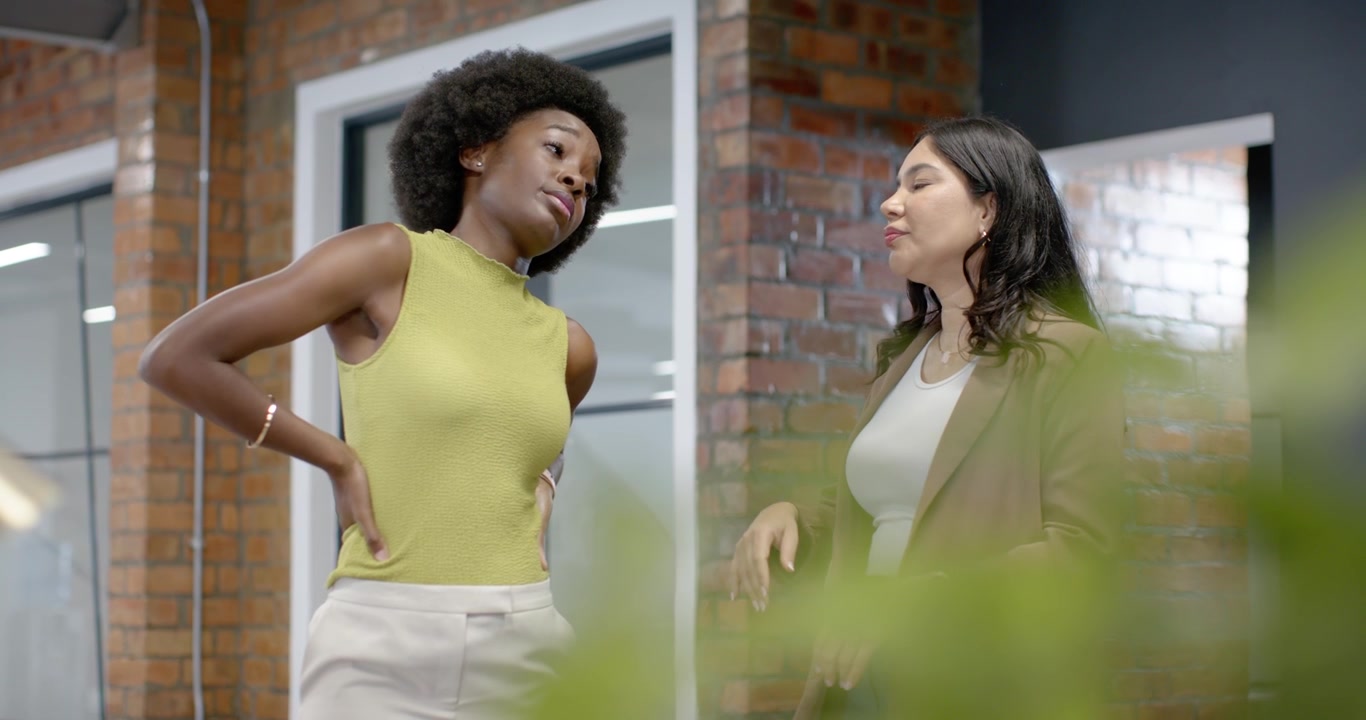 Colegas de trabalho diversas discutindo trabalho no escritório enquanto a câmera se move para a direita, com uma mulher gesticulando perto de uma planta