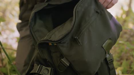 soldier places glove inside two-layer bag, stands up with boot firmly pressing into grass, preparing to march as blurry white flies hover around, capturing outdoor military movement