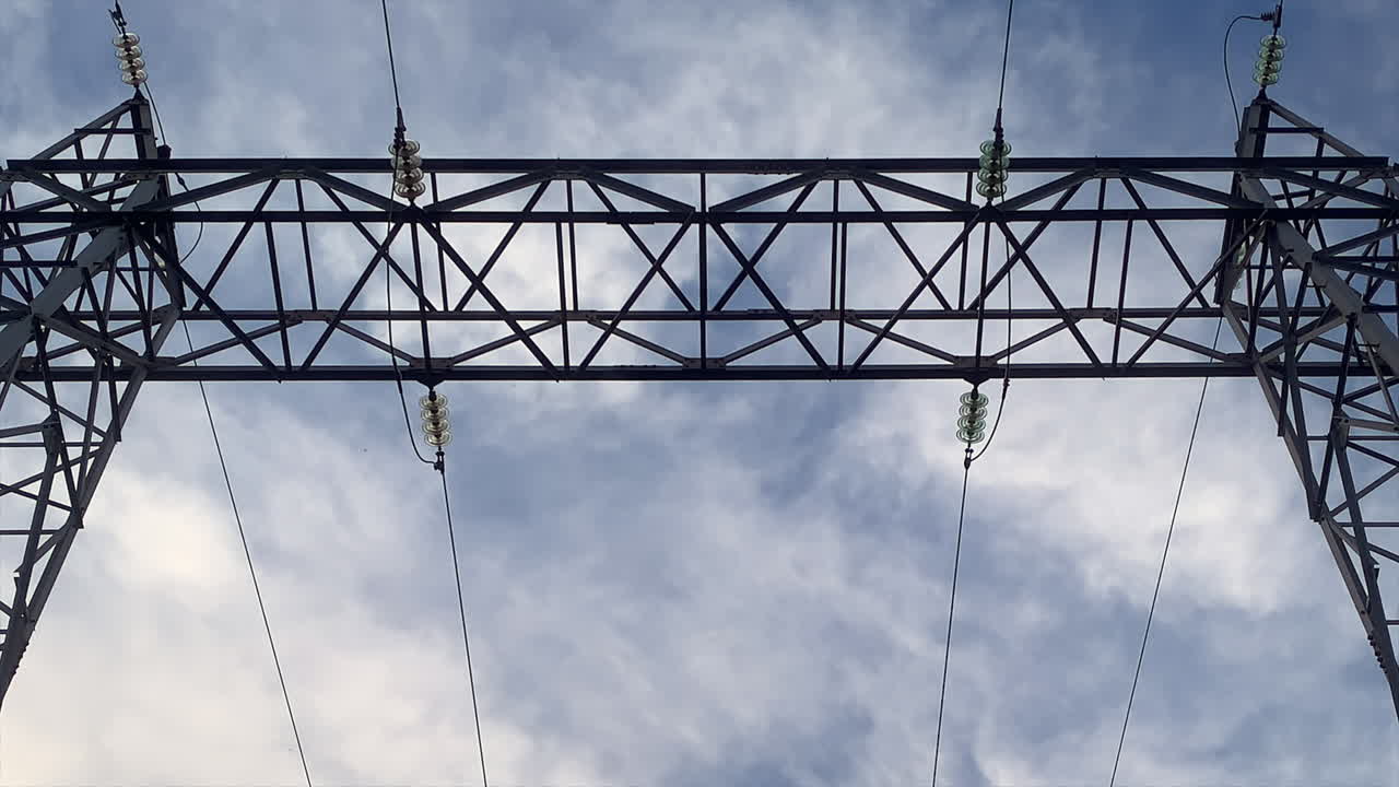 Static upward shot slowly zooming toward a symmetrical high-voltage electricity transmission gantry with power lines and insulators silhouetted against a cloudy sky