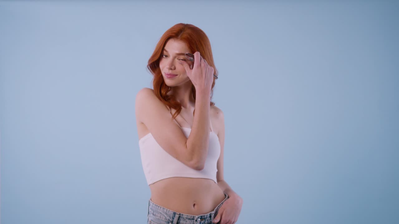 Redhead young woman in white crop top poses with confidence, doing playful gestures while holding a makeup brush in different angles.