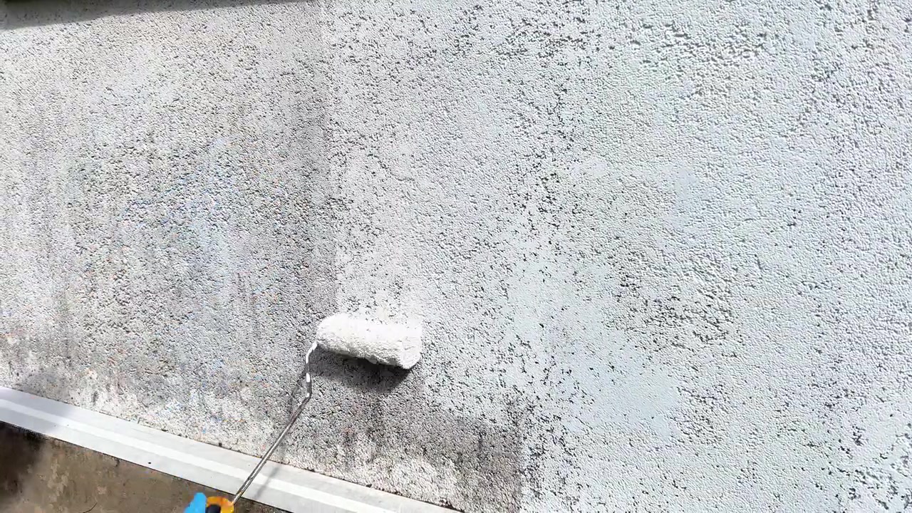 A person in blue protective gloves uses a paint roller to apply fresh white paint to a weathered stucco wall. Perfect for home renovation, exterior painting, and repair videos.