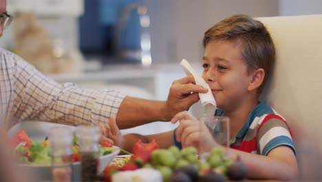 Glücklicher-Kaukasischer-Großvater-Am-Tisch,-Der-Beim-Familienessen-Das-Gesicht-Seines-Enkels-Abwischt