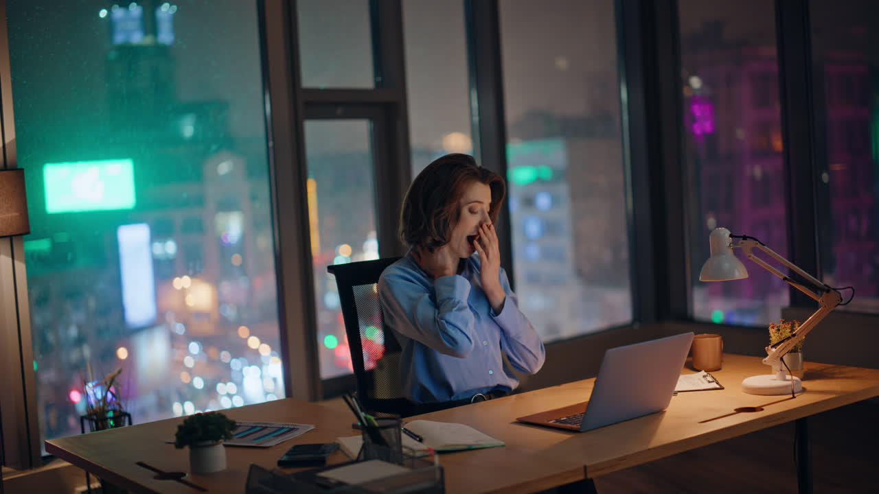 Late evening worker yawning at workplace looking laptop. Tired businesswoman