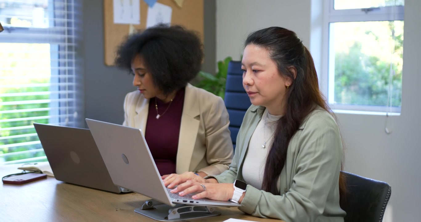 Diverse female coworkers reading then shifting to typing on laptops at office desk to finish report