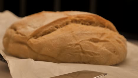cacerola lenta sobre un cuchillo serrado hacia una hogaza de pan de masa agria recién horneada cubierta con harina sentada en un banco de cocina, poca profundidad de campo