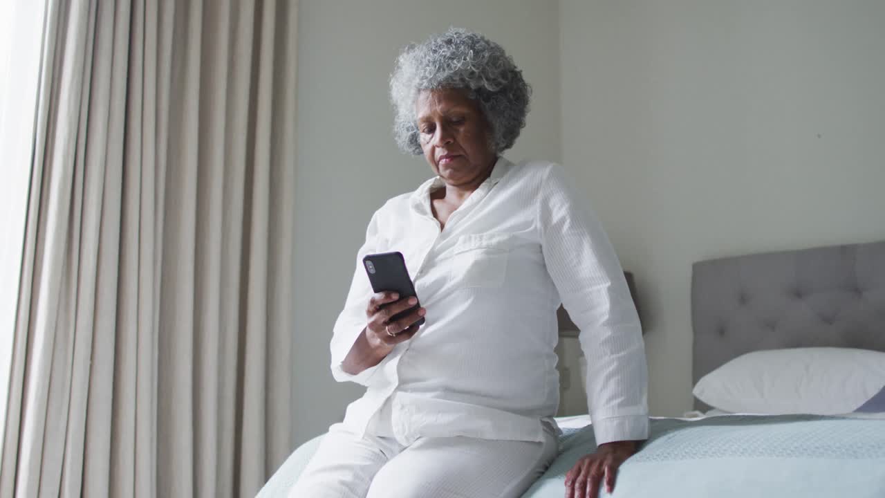 Anziana donna afroamericana che usa lo smartphone mentre è seduta sul letto a casa.