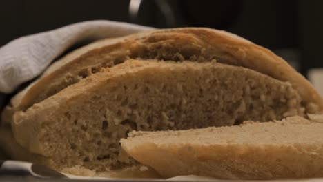 Slow-pan-around-freshly-cut-and-baked-loaf-of-sour-dough-bread-with-piece-sitting-to-side-and-knife-in-foreground-on-kitchen-bench