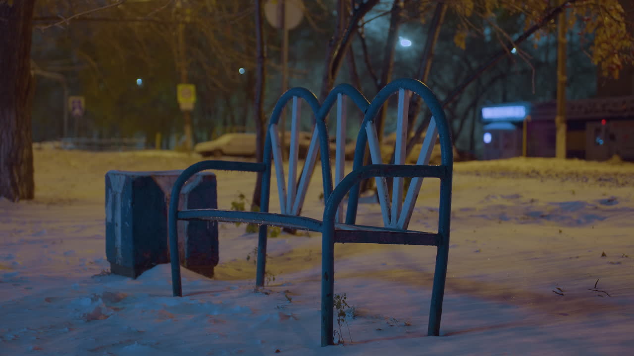 눈으로 인 벤치는 겨울 저녁에 혼자 서 있으며, 부드러운 주변 빛, 주변 나무 및 도시 요소의 흐릿한 배경에 의해 조명됩니다.